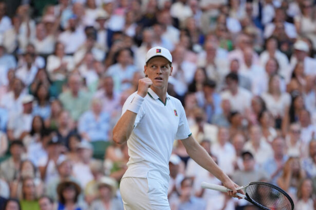 Tennis player in a white outfit and cap fists raised on court, with a large cheering crowd in the stands behind him.