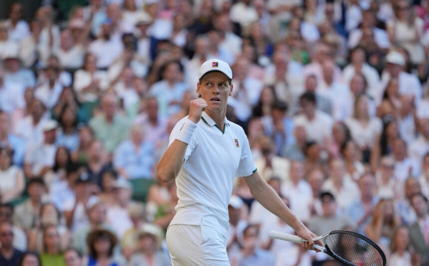 Tennis player in a white outfit and cap fists raised on court, with a large cheering crowd in the stands behind him.