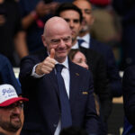 Man in a dark suit and tie giving a thumbs-up at a sports event, with a FIFA badge on jacket and cheering crowd behind him.