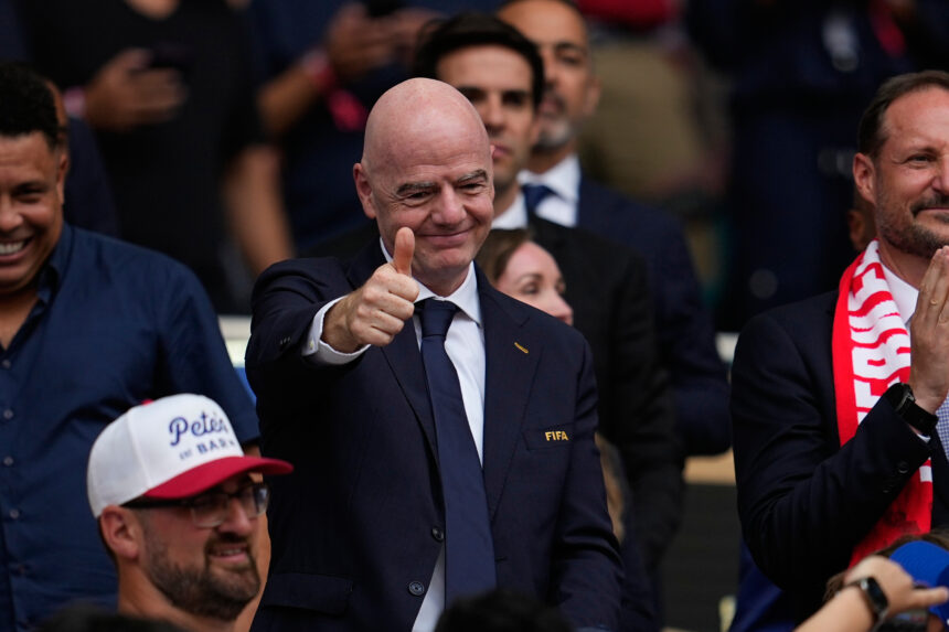 Man in a dark suit and tie giving a thumbs-up at a sports event, with a FIFA badge on jacket and cheering crowd behind him.