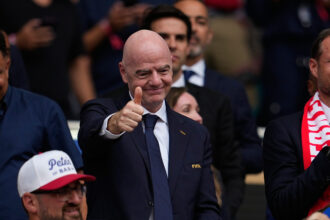 Man in a dark suit and tie giving a thumbs-up at a sports event, with a FIFA badge on jacket and cheering crowd behind him.