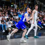 Basketball players sprinting on a crowded indoor court, one in blue driving forward while a defender in white watches nearby.