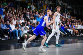 Basketball players sprinting on a crowded indoor court, one in blue driving forward while a defender in white watches nearby.