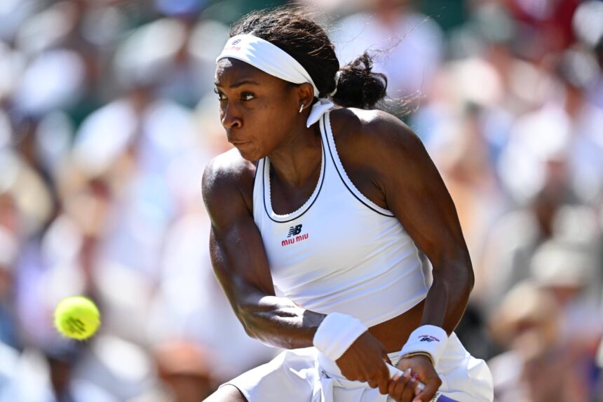 Female tennis player in a white sleeveless outfit and headband mid-swing on a sunny court with a blurred crowd in the background, focused on the ball.