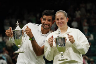 Two athletes in white outfits proudly hold large silver trophies on a podium, smiling after a victory in front of a cheering crowd.