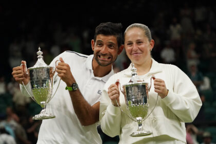 Two athletes in white outfits proudly hold large silver trophies on a podium, smiling after a victory in front of a cheering crowd.