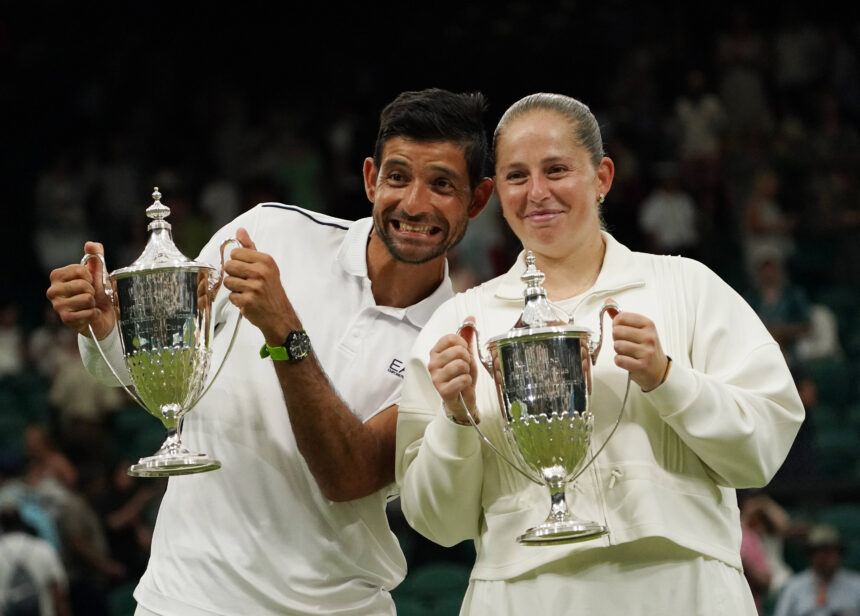 Two athletes in white outfits proudly hold large silver trophies on a podium, smiling after a victory in front of a cheering crowd.