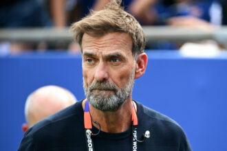 Close-up of a middle-aged man with a gray beard and dark shirt, wearing a lanyard, against a blue background.