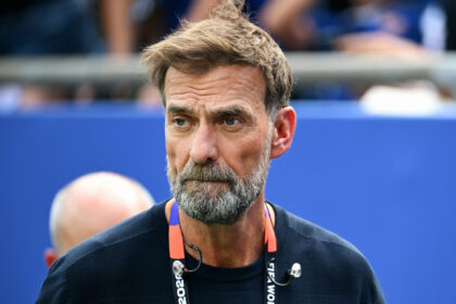 Close-up of a middle-aged man with a gray beard and dark shirt, wearing a lanyard, against a blue background.