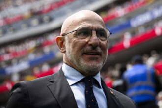 Close-up portrait of a smiling man in a suit and round glasses at a stadium with blurred red and blue seating in the background.