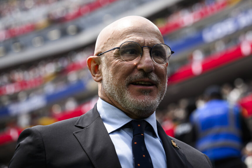 Close-up portrait of a smiling man in a suit and round glasses at a stadium with blurred red and blue seating in the background.