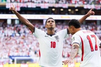 England footballers celebrate a goal with arms raised as a full stadium cheers behind them.