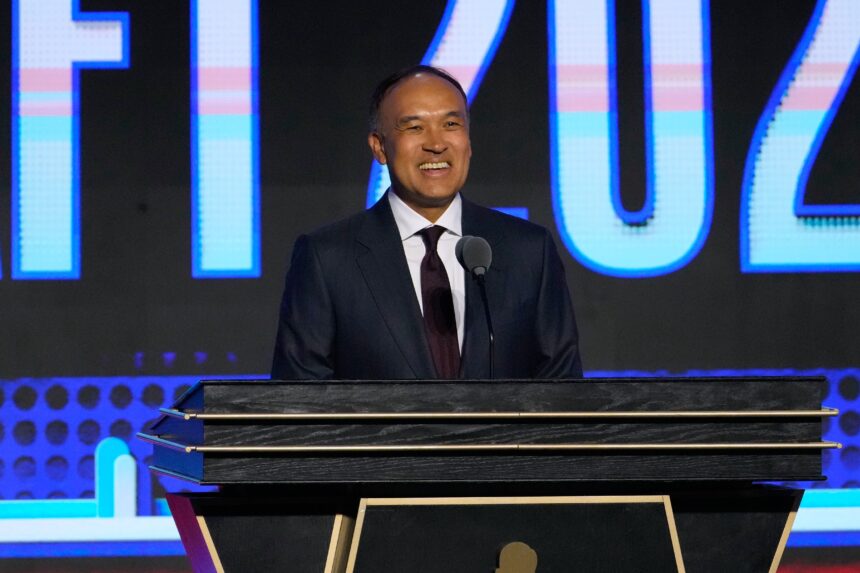 Man in a dark suit smiles while speaking at a podium on a stage with a blue, lit backdrop behind him.