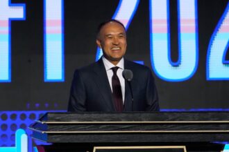 Man in a dark suit smiles while speaking at a podium on a stage with a blue, lit backdrop behind him.