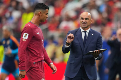 Soccer coach in a navy suit speaks to a player in a red jersey on the field, gesturing with his hand.