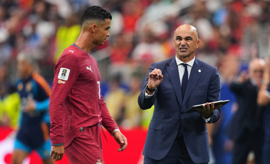 Soccer coach in a navy suit speaks to a player in a red jersey on the field, gesturing with his hand.