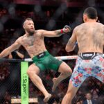 Two male UFC fighters collide in the octagon as the left fighter throws a punch toward his tattooed opponent amidst a crowded arena.